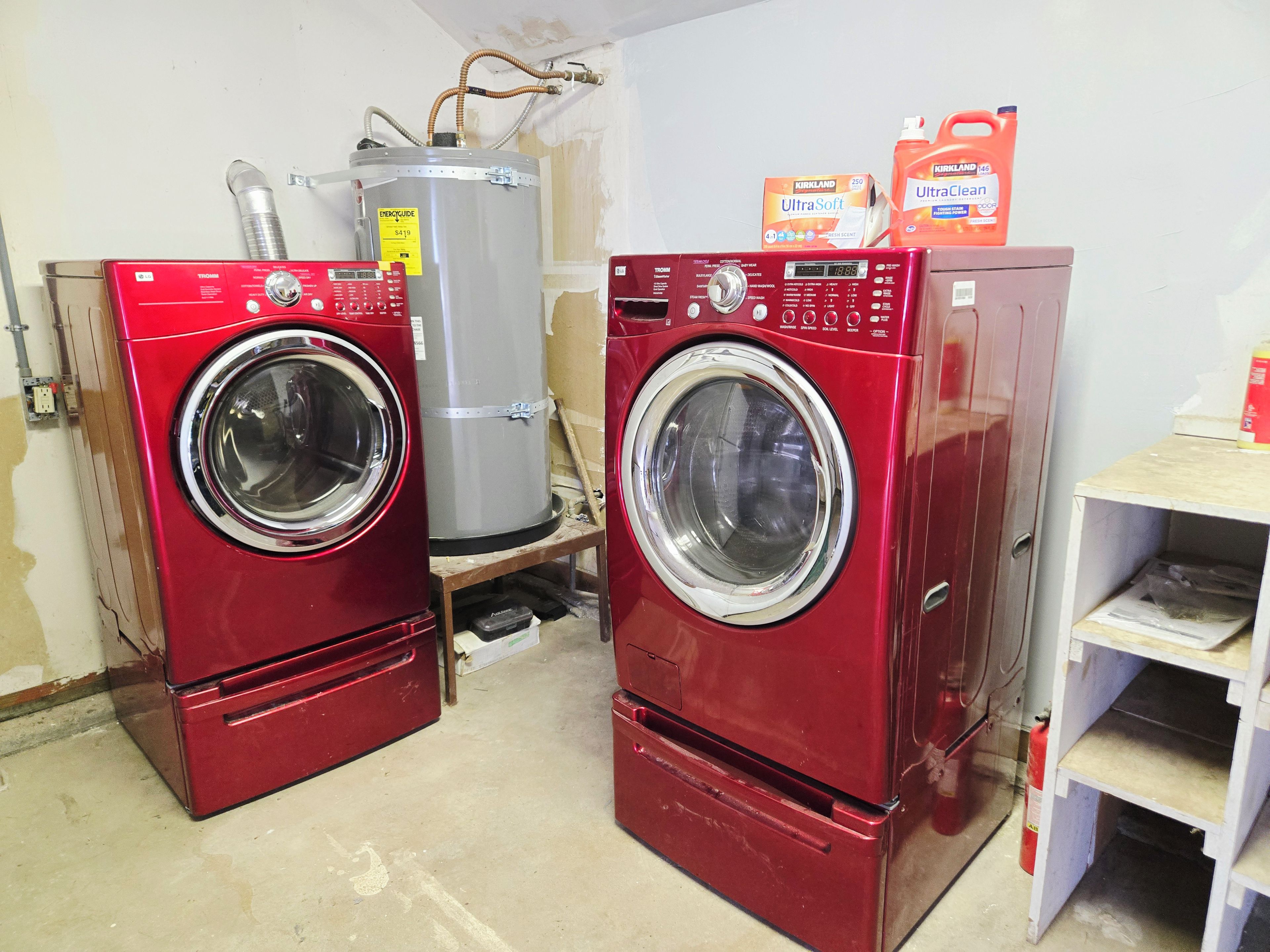 Dos lavadoras rojas de carga frontal, un calentador de agua grande y botellas de detergente en una habitación con paredes sin terminar.