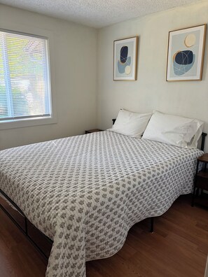 Un dormitorio con cama, ventana con persianas y dos cuadros abstractos en la pared.