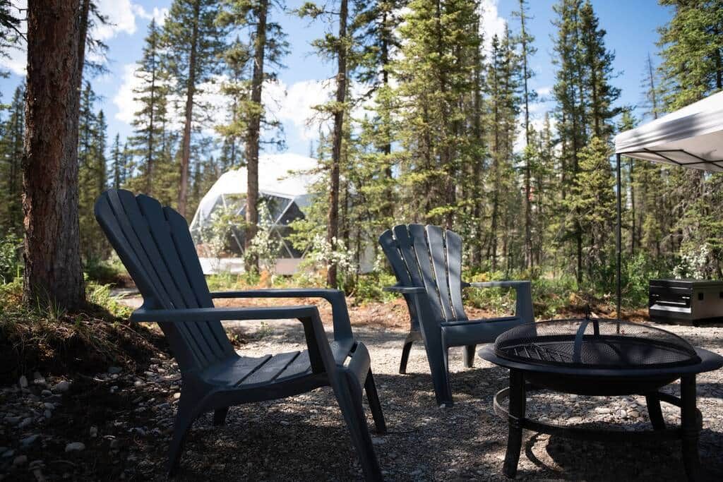 Deux fauteuils Adirondack, un foyer et une tente, le tout installé dans une zone boisée.