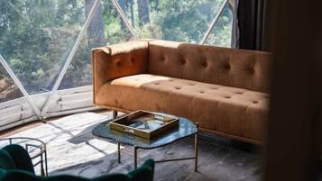A room with a brown sofa, a glass coffee table, and a view of a forest through a large window.