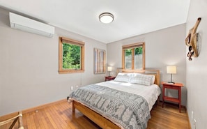 Ein Schlafzimmer mit einem hölzernen Bett, zwei Nachttischen, einer Wand-Klimaanlage und einem Fenster mit Blick auf Grünflächen.