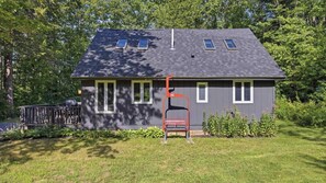 A two-story house with a dark roof, multiple windows, and a red chair in the front yard. - Cozy 2-bedroom chalet in delightful Gunstock Acres (Gilford)