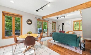 A modern living room with a dining table, chairs, a sofa, and large windows. - Cozy 2-bedroom chalet in delightful Gunstock Acres (Gilford)