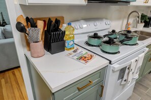 Eine Küche mit weißer Arbeitsplatte, einem Herd und einem Topfset. Auf der Arbeitsplatte steht eine Flasche Öl und ein Reibe.