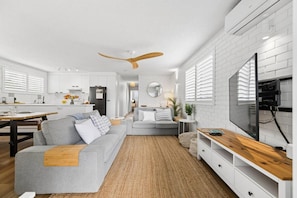 A modern kitchen with white cabinetry, a dining area with a wooden table, a grey sofa, and a TV on a wooden stand. - Muroy - Beachside Apartment @ Cotton Tree (Maroochydore)