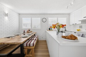 A modern kitchen with white cabinetry, a wooden dining table, and a bench. - Muroy - Beachside Apartment @ Cotton Tree (Maroochydore)