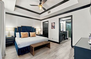 Een moderne slaapkamer met een groot bed, nachtkastjes, een bankje, een plafondventilator en een badkamer met spiegel en wastafel.