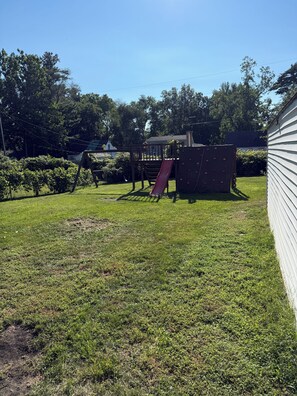 A backyard with a play structure, a red slide, and a white house in the background. - Boho Bungalow walking distance from Notre Dame (South Bend)