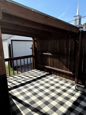 A wooden deck with a checkered floor, a metal railing, and a view of a white garage door and a church steeple in the background. - Boho Bungalow walking distance from Notre Dame (South Bend)