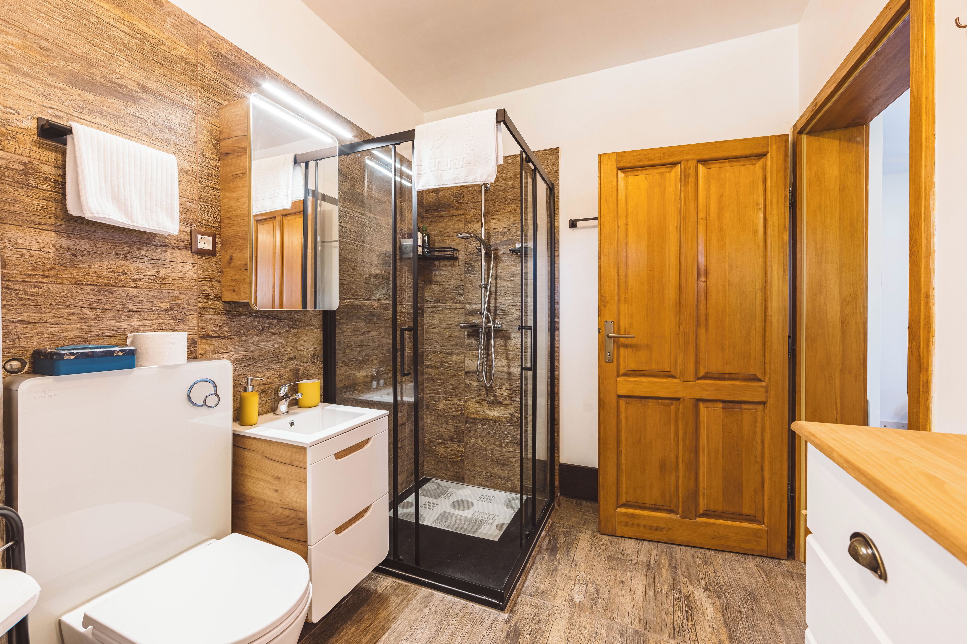 A modern bathroom with a glass shower enclosure, wooden wall paneling, a white toilet, and a wooden door.