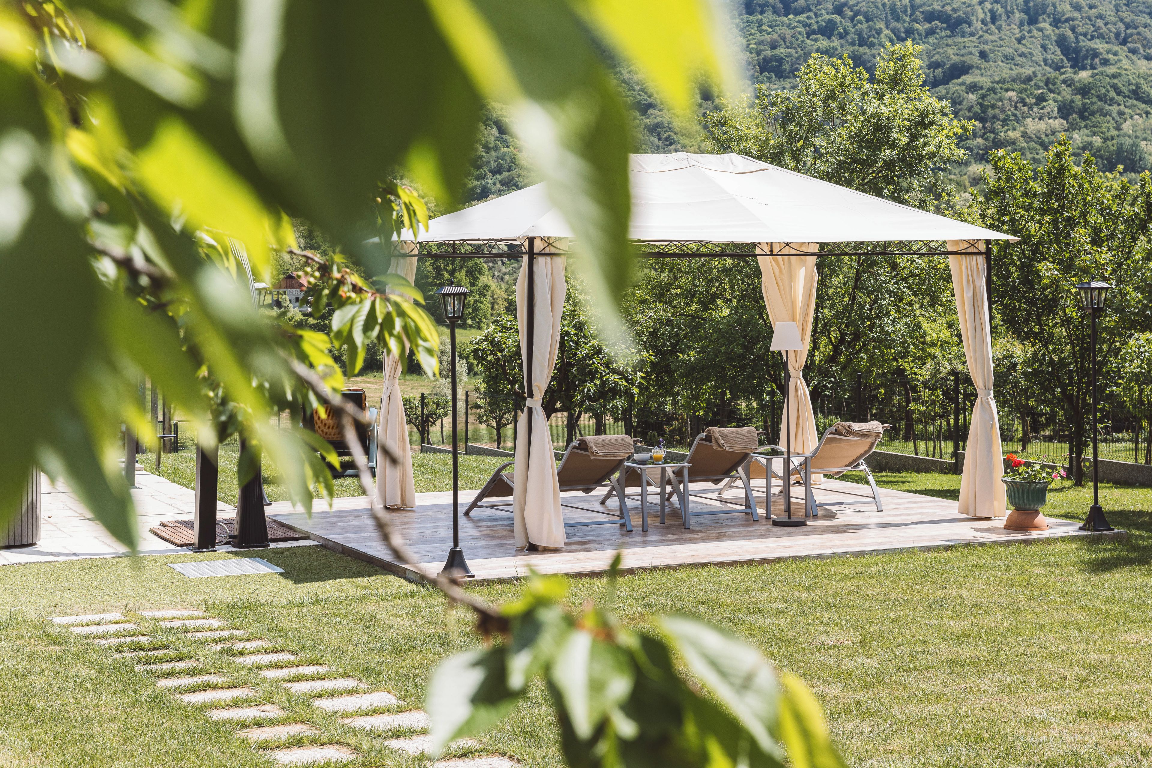 A gazebo with four chairs and a table, surrounded by a grassy area with trees in the background.