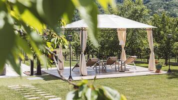 A gazebo with four chairs and a table, surrounded by a grassy area with trees in the background.