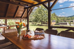 Un'area pranzo all'aperto con un tavolo di legno, sedie, una bottiglia di vino, un bicchiere, un cesto di pane e vista su un paesaggio verde con alberi e colline.