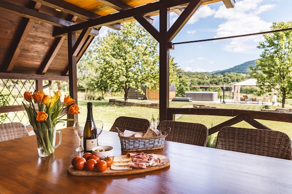 Un'area pranzo all'aperto con un tavolo di legno apparecchiato per un pasto, con una bottiglia di vino, un bicchiere, un cestino di pane, pomodori e formaggio.