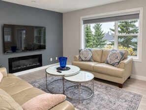 A living room with a beige sofa, a glass coffee table, a fireplace, and a large window with a view of trees. - Panorama Hills Manor | Private Luxury Main & Upper | Near YYC, Easy Banff Access (Calgary)