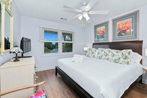 Un dormitorio con cama, ventilador de techo, ventana con persianas y dos cuadros enmarcados con motivos florales en la pared.