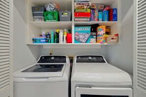A laundry room with two washing machines and shelves above containing various board games and cleaning supplies. - 3 Ocean Park Court, Isle of Palms (Isle of Palms)