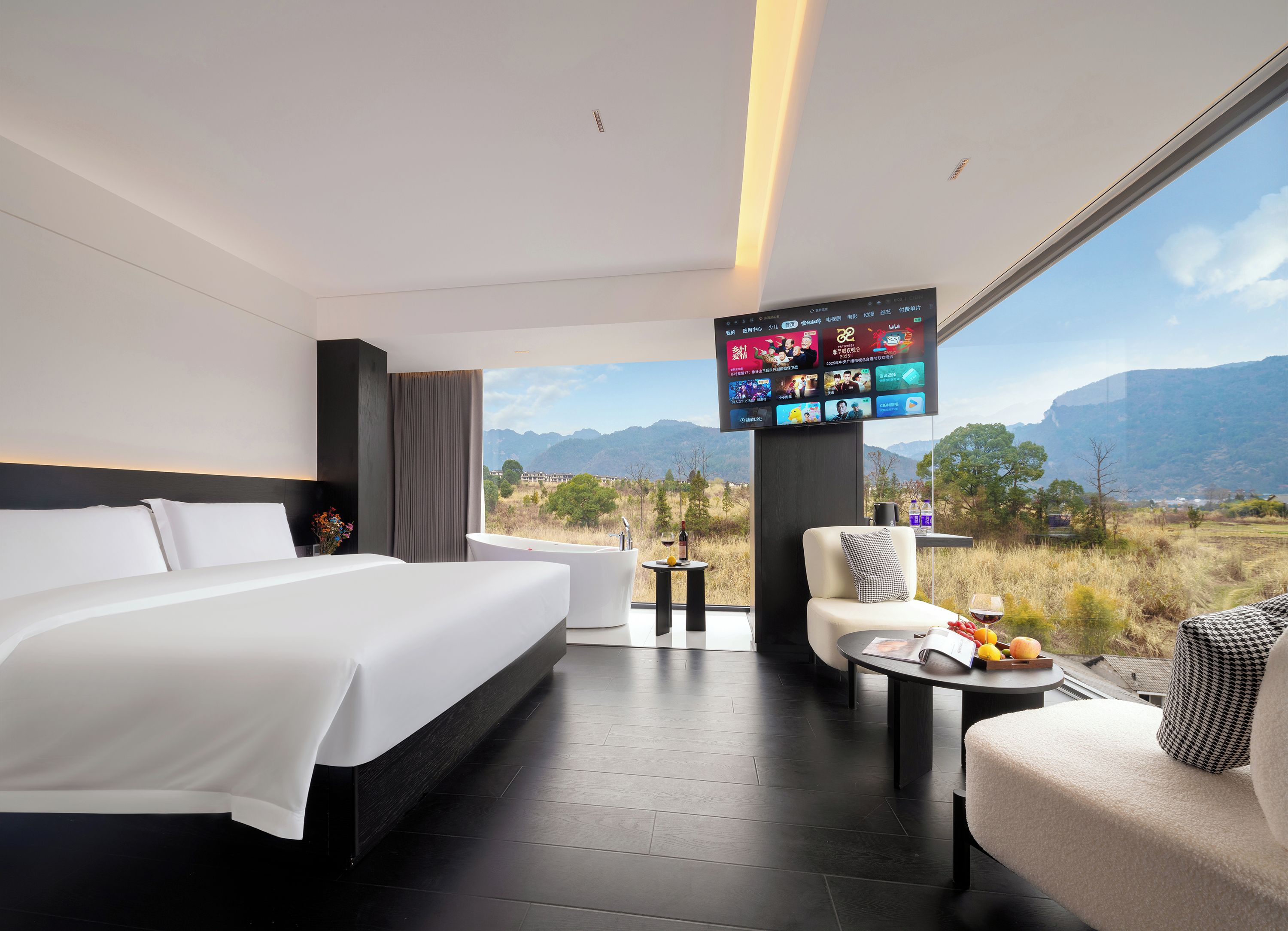 A modern hotel room with a large bed, a seating area with a view, and a flat-screen TV mounted on the wall.