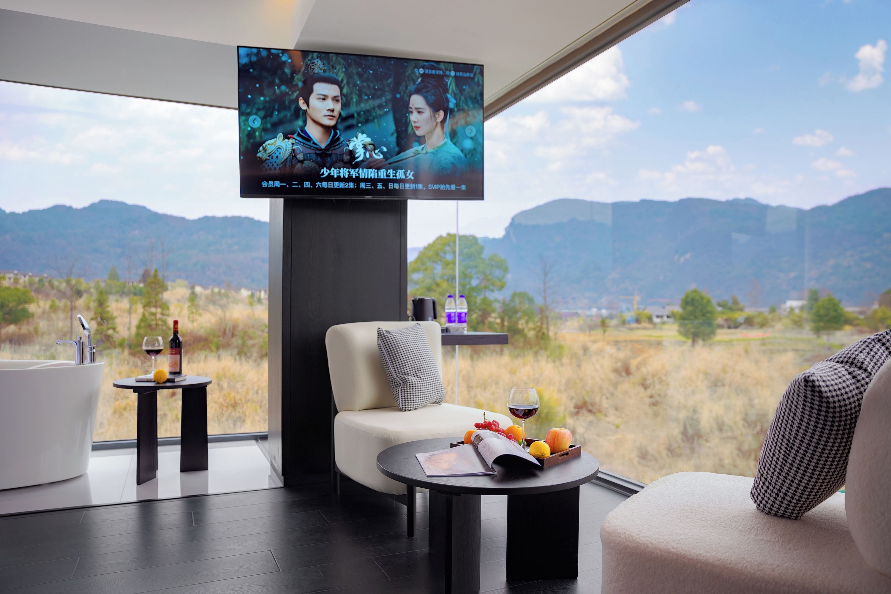 A modern room with a large TV mounted on the wall, a sofa, a coffee table with fruits and a wine glass, and a view of mountains through large windows.