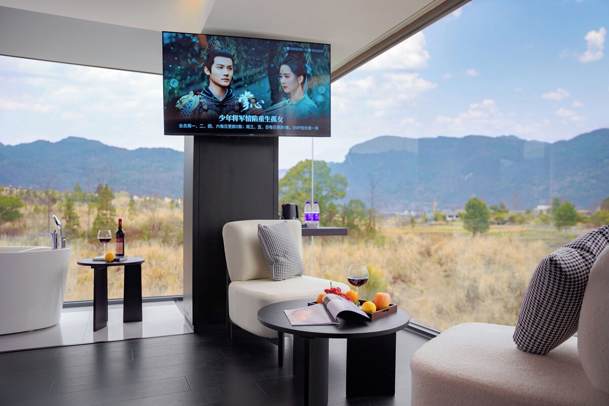 Ein modernes Zimmer mit einem großen an der Wand befestigten Fernseher, einer Couch, einem Couchtisch mit Früchten und einem Weinglas, und Blick auf Berge durch große Fenster.
