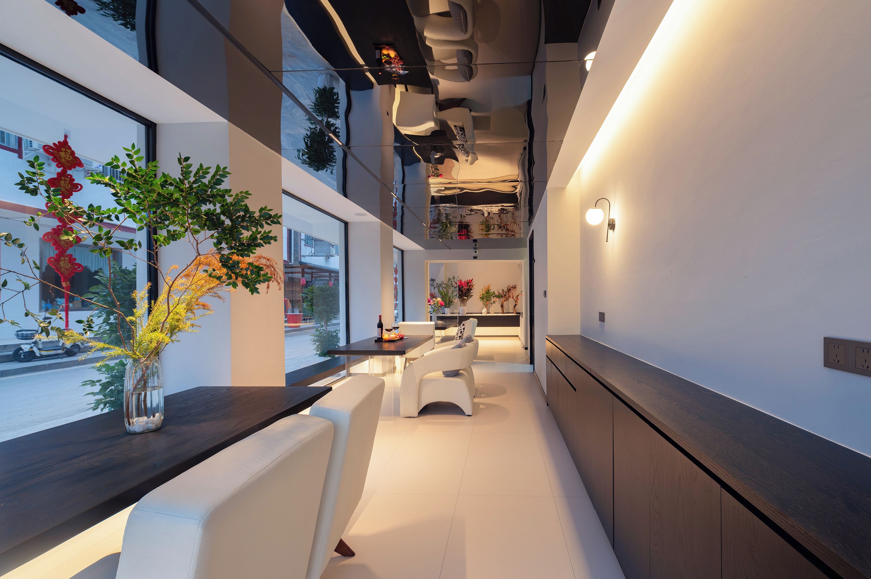 A modern interior with a long dining table, white cushioned seating, and a view through large windows.