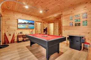 A pool table with a red felt surface, a television mounted on the wall, and a billiards-themed wall decoration. - Modern Rustic Cabin - Hot tub, Shuffleboard, Pool Table Nestled in Trees (Logan)