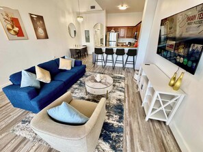 A modern living room with a blue sofa, a beige armchair, and a coffee table. There is a kitchen area with a bar and stools in the background. - Balcony Lake Views | Pool, Gym, WiFi & Parking (Orlando)