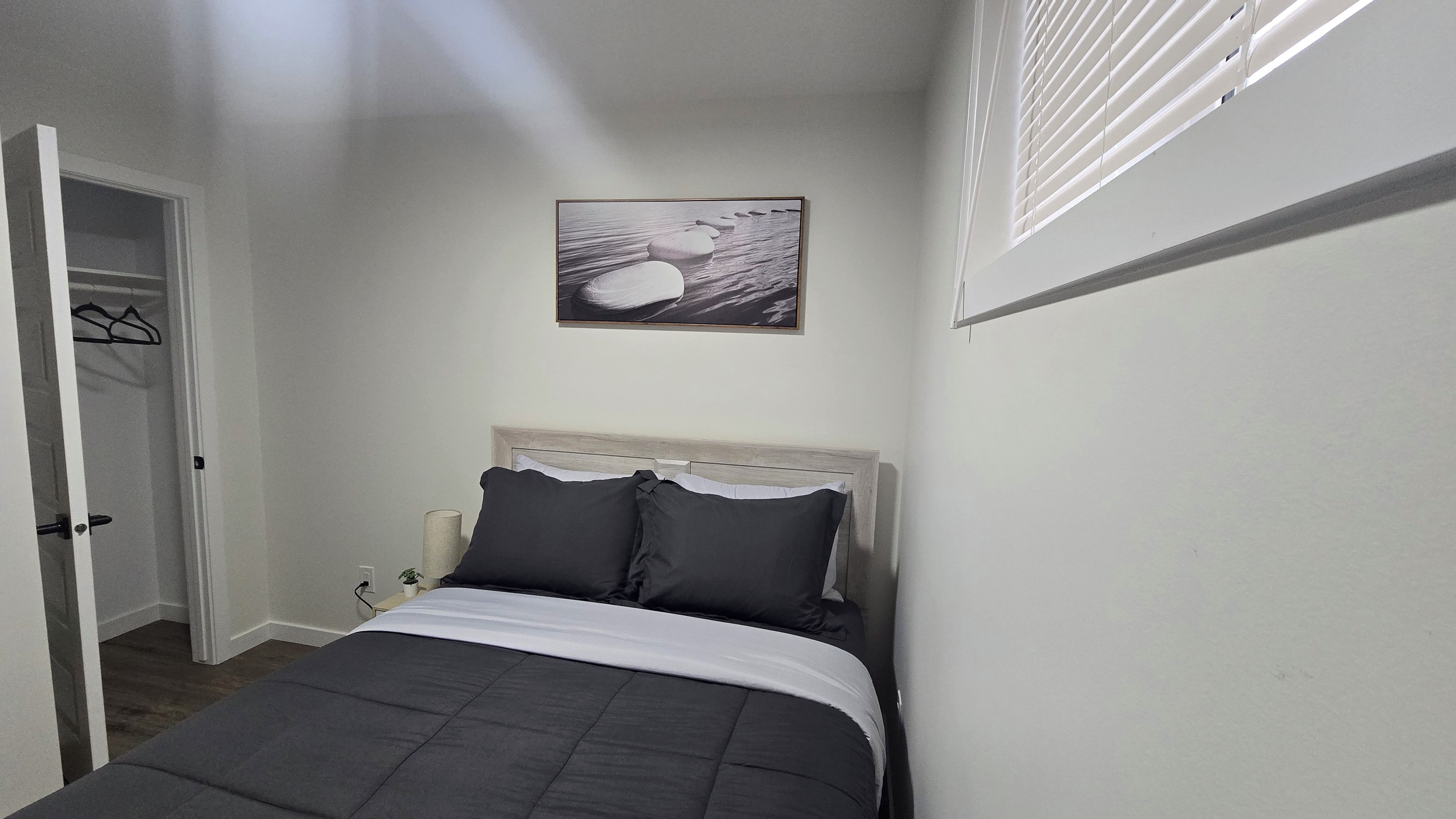 Ein Schlafzimmer mit Bett, Tür, einem an der Wand hängenden Bild und einem Fenster mit Jalousien.