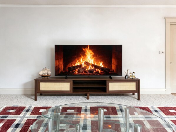 Un televisor montado sobre un soporte de madera con una chimenea a la vista, una mesa de centro de vidrio y una alfombra estampada.