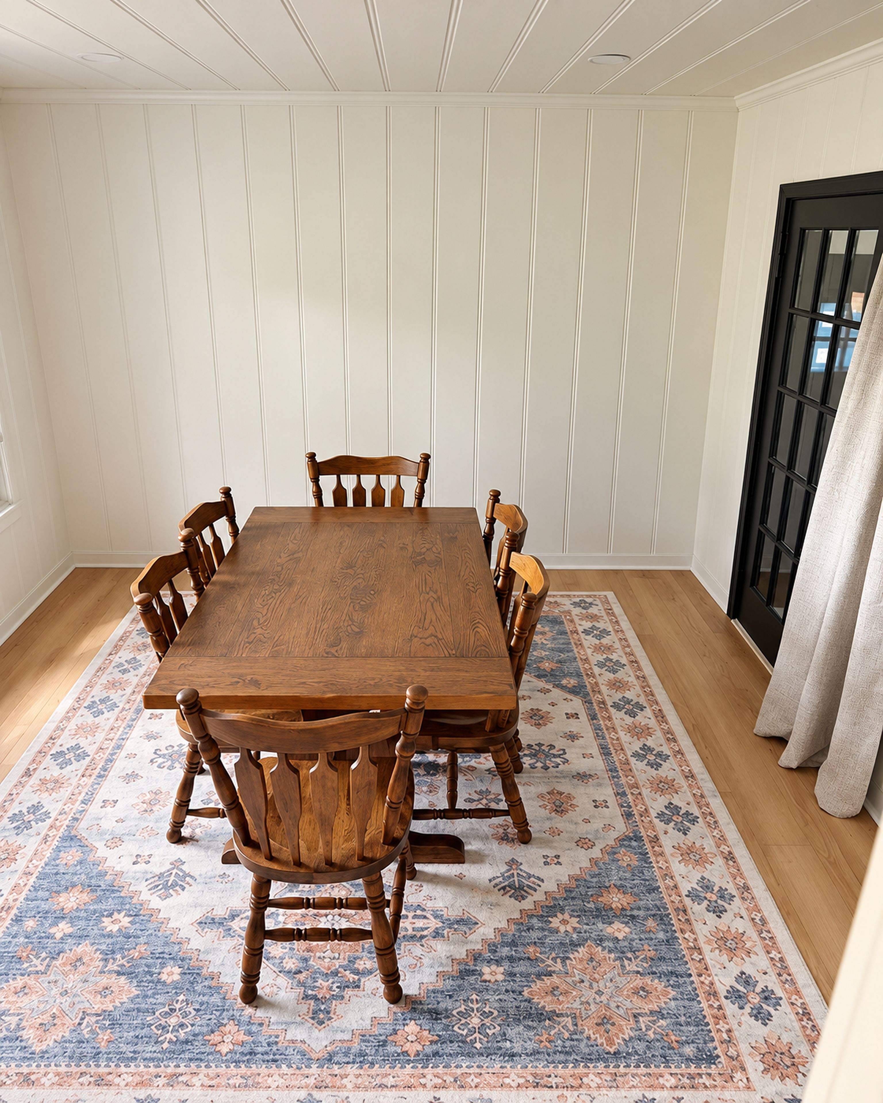 A dining room with a wooden table and chairs, a patterned rug, and white paneled walls.