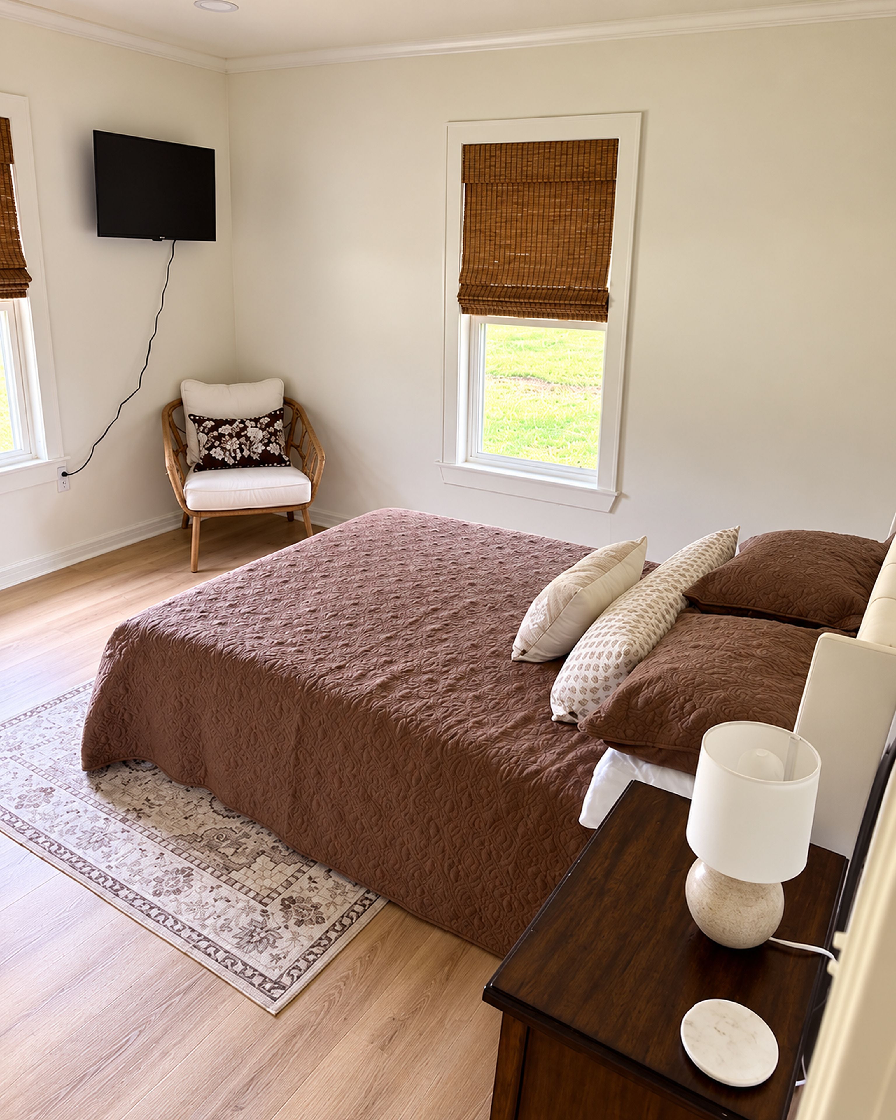 A bedroom with a large bed, a bedside table with a lamp, a chair, a TV mounted on the wall, and a window with a view of the outdoors.