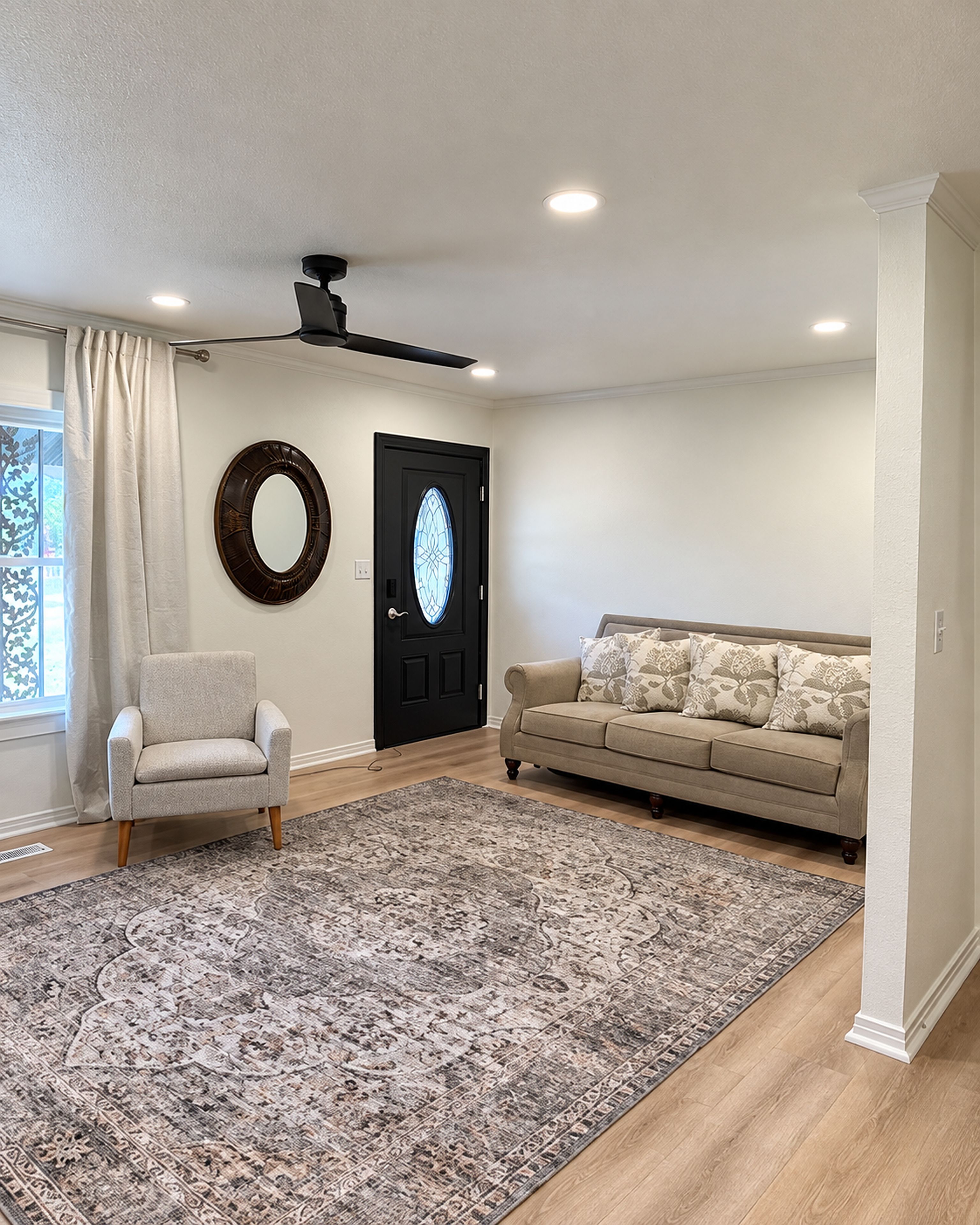 A spacious living room with a large rug, a sofa, a chair, a ceiling fan, and a door with a decorative glass panel.