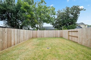 A fenced backyard with a grassy area and trees in the background. - New 2026 4BR Home | Garage | Pets OK | 10 Min to Med Center, NRG & Downtown (Houston)