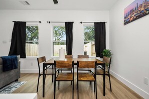 A dining area with a table and chairs, large windows with curtains, a cityscape painting, and a grey sofa. - New 2026 4BR Home | Garage | Pets OK | 10 Min to Med Center, NRG & Downtown (Houston)