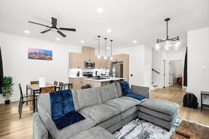 A modern living room with a grey sectional sofa, a dining area with a table and chairs, a kitchen with wooden cabinets, and a ceiling fan. - New 2026 4BR Home | Garage | Pets OK | 10 Min to Med Center, NRG & Downtown (Houston)
