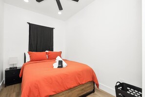 A bedroom with a bed, black curtains, a nightstand, a lamp, and a black basket. - New 2026 4BR Home | Garage | Pets OK | 10 Min to Med Center, NRG & Downtown (Houston)