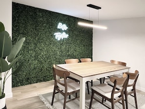 A dining area with a wooden table and chairs, a green wall with a neon sign, and a hanging light fixture. - Westmount Retreat | 5 Bedrooms, 2 Baths & Free Parking (Hamilton)
