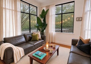 A modern living room with a brown leather sofa, a coffee table with books and candles, and large windows with curtains. - Sol Villa Forest Retreat & Walk to Town (Margaret River)