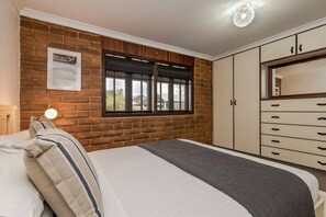 A bedroom with a brick wall, a bed with pillows, a window with blinds, a wardrobe, and a bedside lamp. - Le Souef Authentic MR Retreat With Cosy Fire (Margaret River)