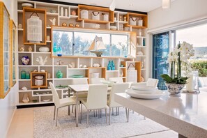 Een moderne eethoek met een groot raam, witte meubels en decoratieve objecten op planken.