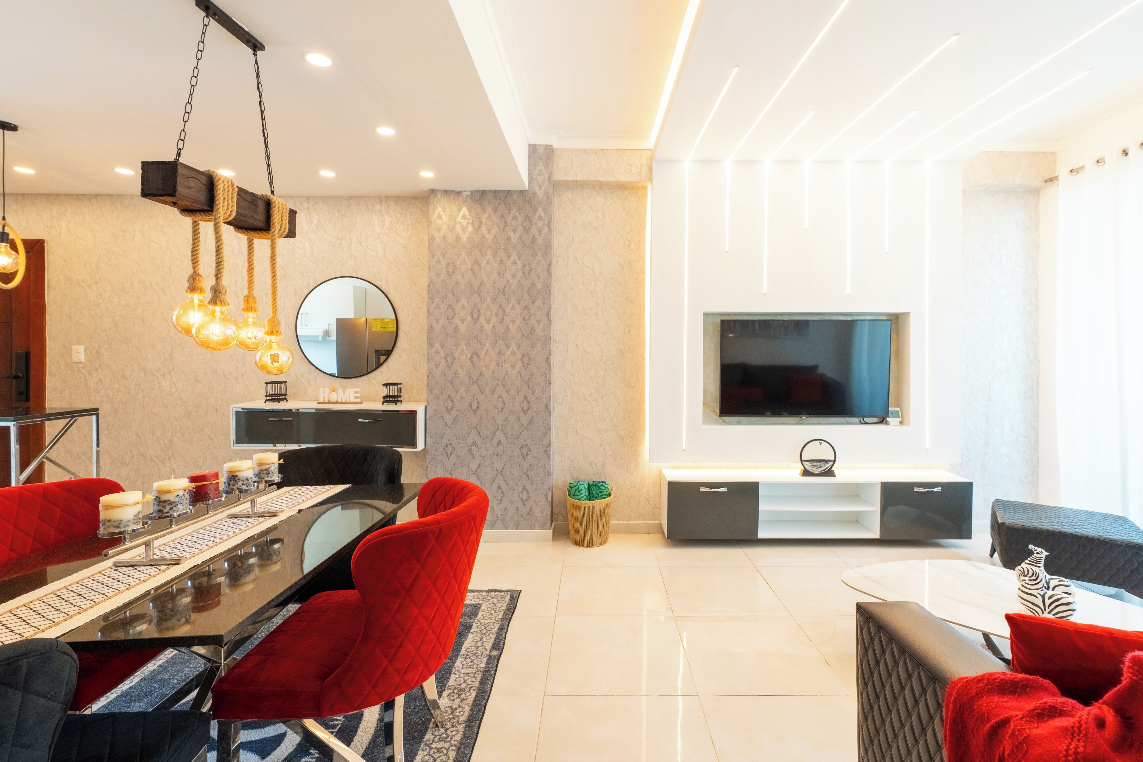A modern living room with a dining area, a flat-screen TV mounted on the wall, and a chandelier.