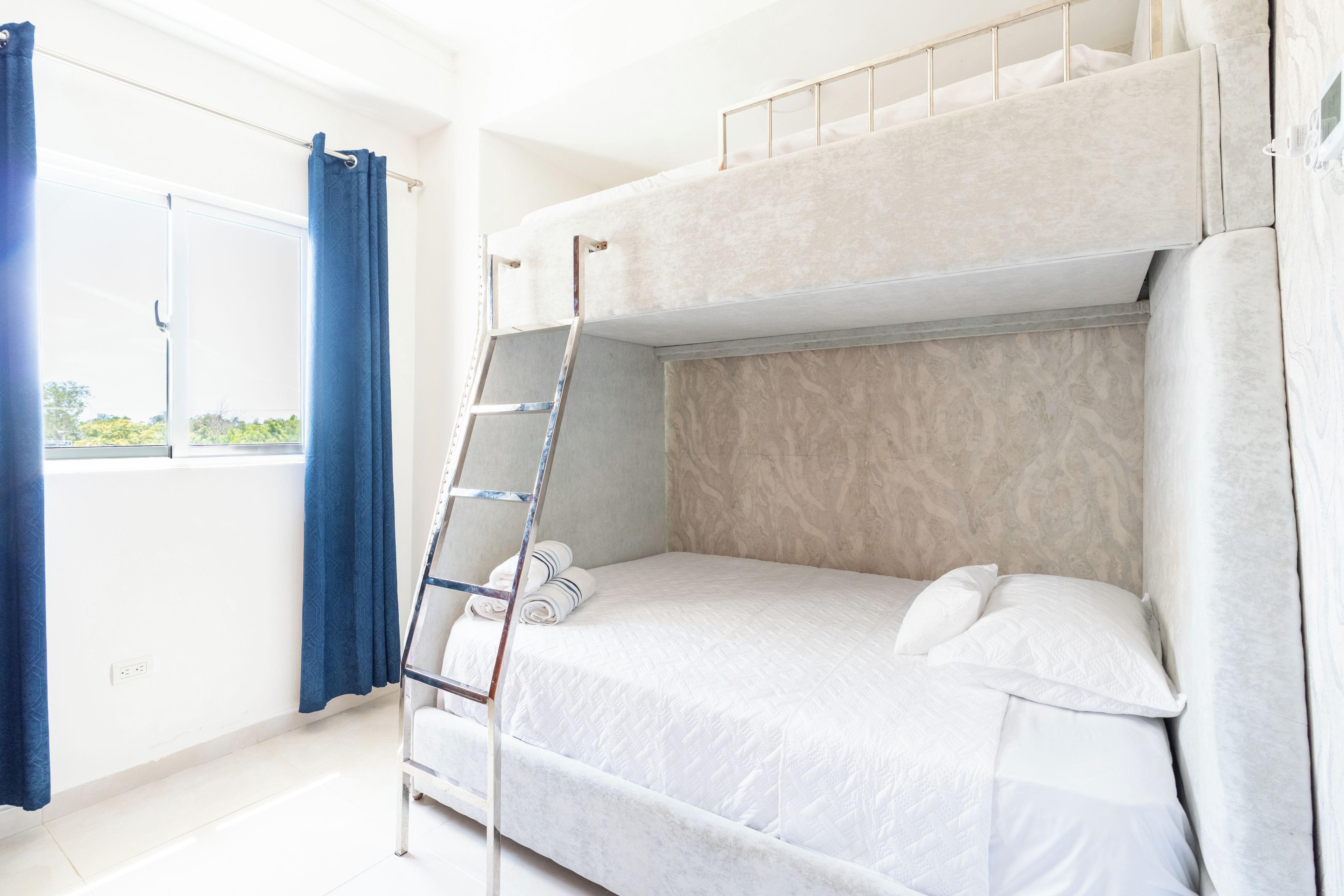 A bunk bed room with a ladder, a window with curtains, and a view of greenery outside.
