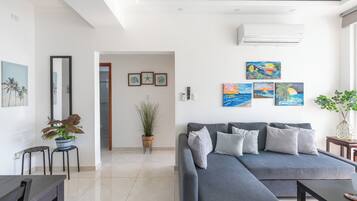 Sala de estar moderna com sofá seccional cinza, mesa de centro e mesa de jantar. Há quadros nas paredes, um vaso com planta e um espelho grande.
