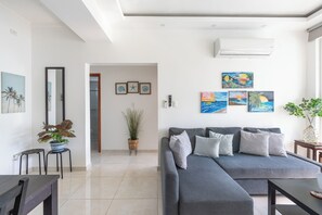 Sala de estar moderna com sofá seccional cinza, mesa de centro e mesa de jantar. Há quadros nas paredes, um vaso com planta e um espelho grande.