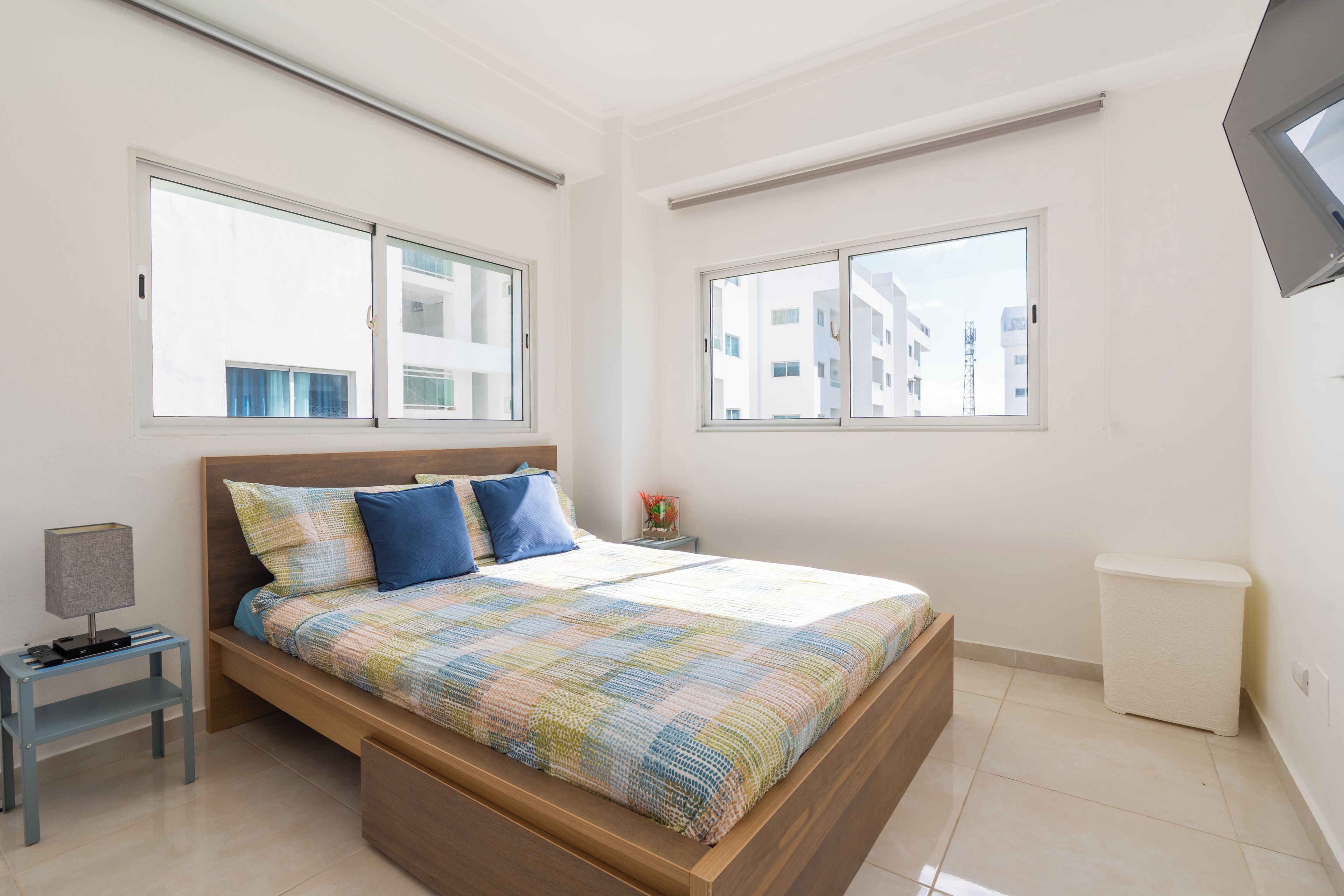 A bedroom with a bed, a nightstand, a lamp, and a view of buildings through the windows.