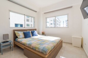 Quarto com cama, mesa de cabeceira, luminária e vista para edifícios pelas janelas.