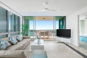 A modern living room with a sofa, coffee table, and TV, a balcony with outdoor furniture, and a large window. - Coastal Heights | Broadwater Views (Southport)