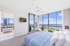 Une chambre avec un grand lit, un ventilateur de plafond et une vue sur une étendue d’eau grâce à de grandes fenêtres.