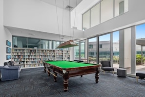 A billiards table in a modern indoor space with a large bookshelf and a view through large windows. - Coastal Heights | Broadwater Views (Southport)