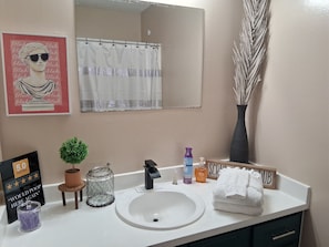 A bathroom with a white sink, a mirror, a shower curtain, a potted plant, a soap dispenser, a towel, and a decorative sign.